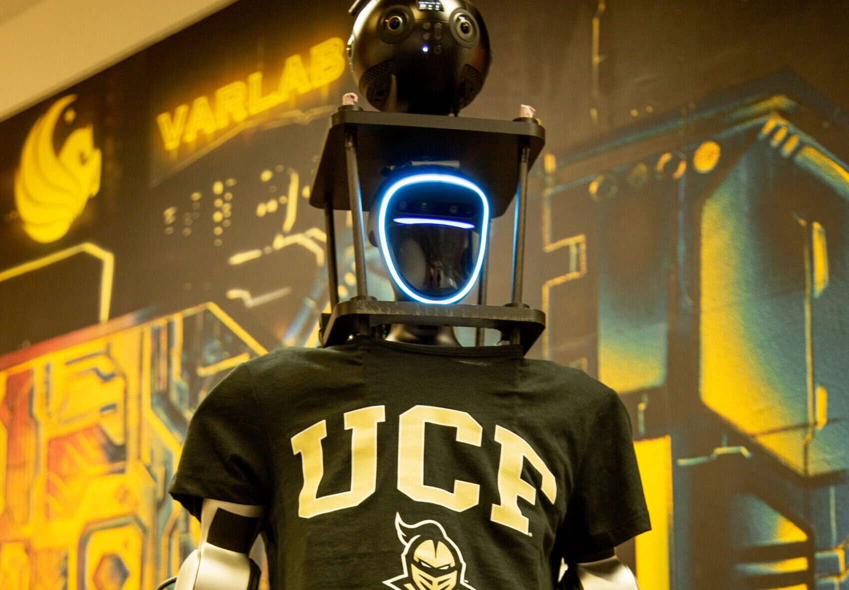 A humanoid robot with a camera mounted on its head, wearing a UCF branded shirt.