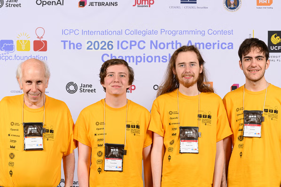 Four men in matching yellow shirts standing in front of a background reading The 2026 ICPC North America Championship.