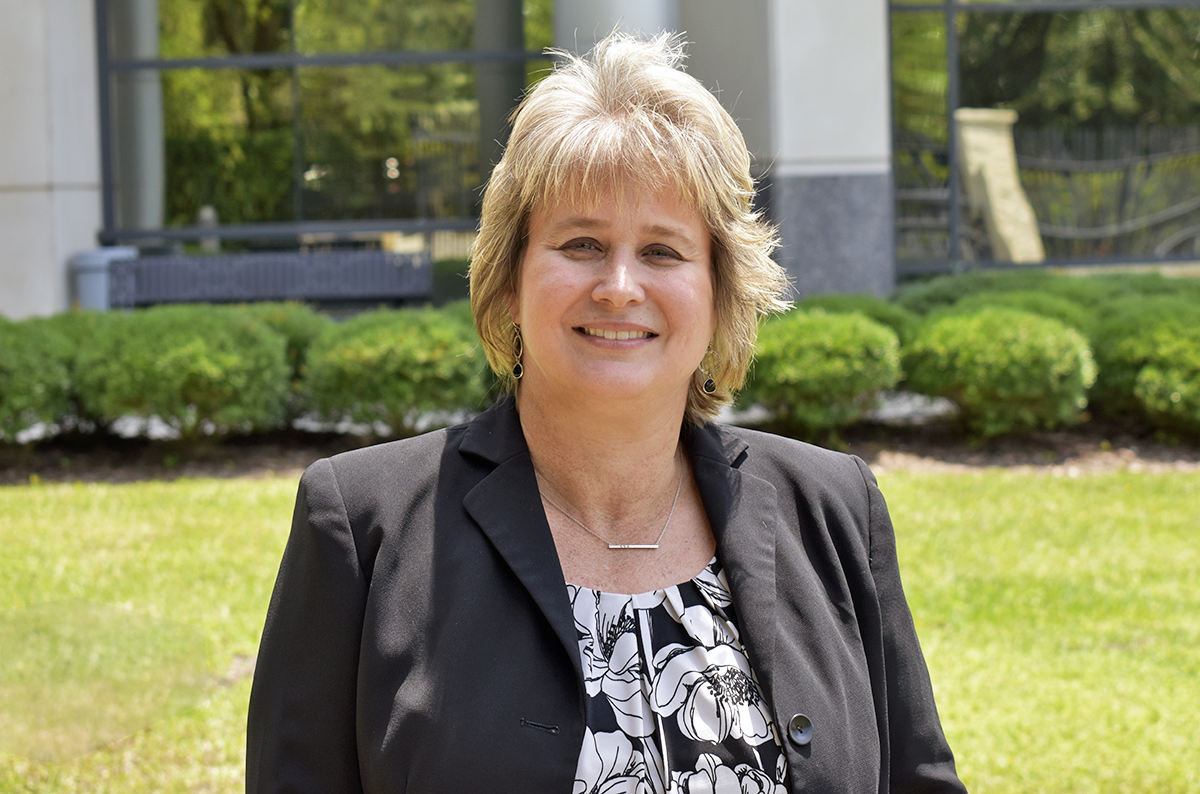 A woman wearing a black suit jacket standing outside in a field with a building behind her.