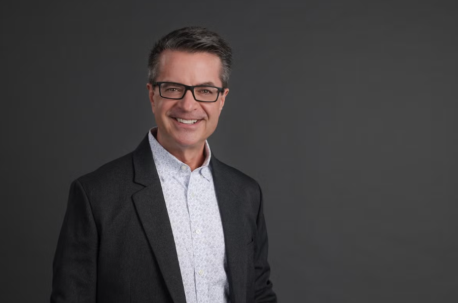 Headshot of a white man wearing glasses and a suit against a gray background.